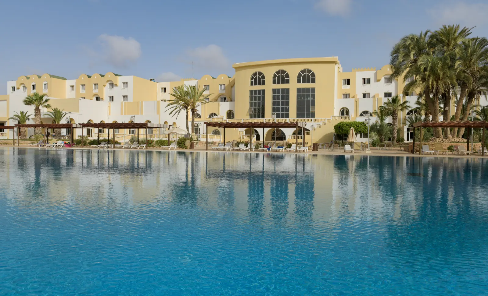 Grande piscine extérieure lagon entourée de palmiers et accès direct à 30 mètres de la plage privée d'Aghir à l'Hôtel Djerba Castille